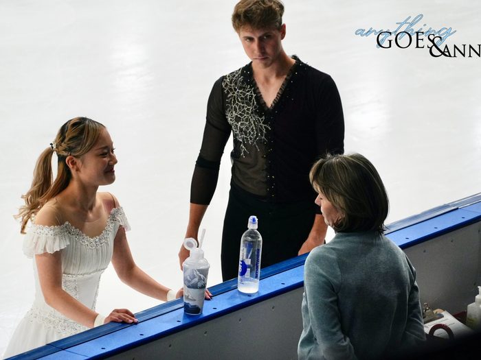 Two skaters stand at the boards speaking to their coach. Miyu Yunoki is smiling broadly at coach Natalia Mishkutenok